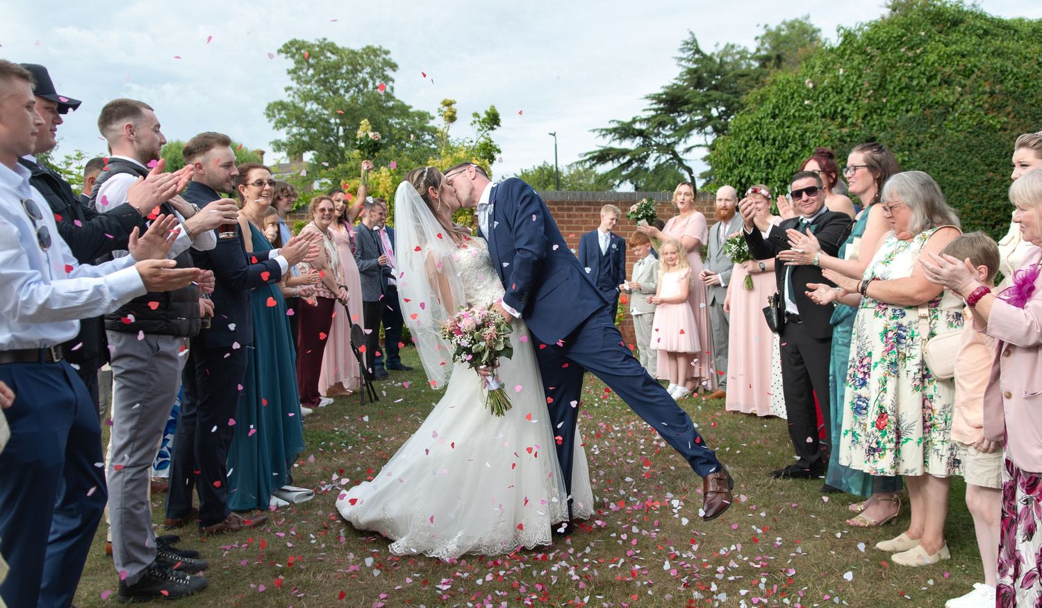 Bride and groom share a kiss while wedding guests throw colorful confetti outdoors at a joyful celebration.