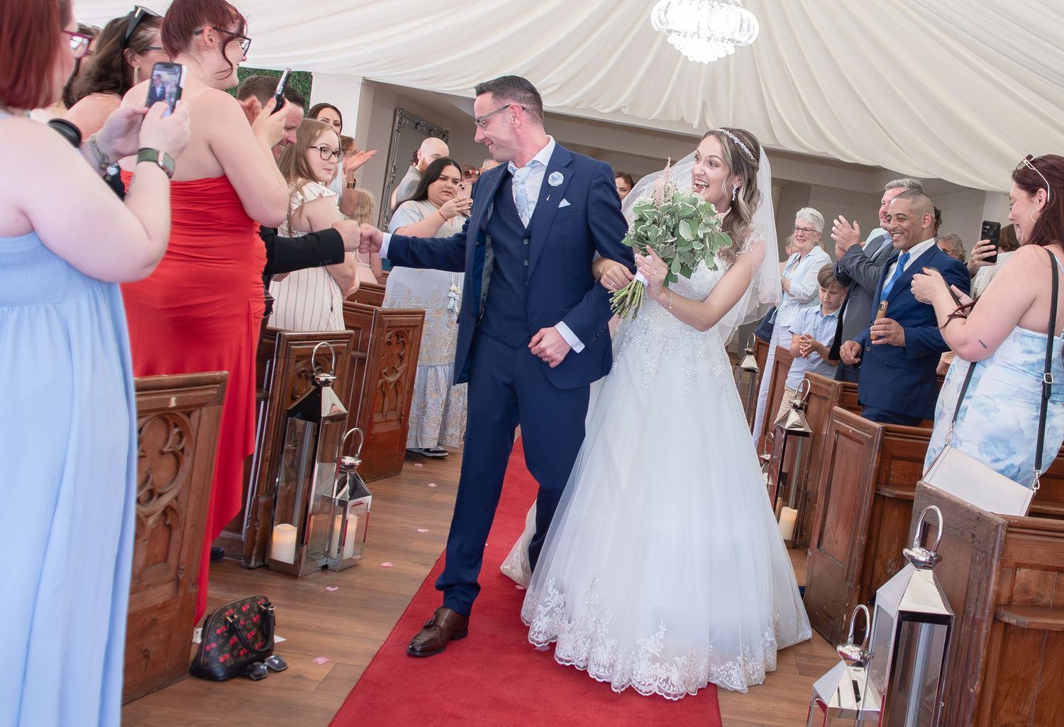 Bride and groom walking down the aisle after wedding ceremony, guests celebrating in church with red carpet.