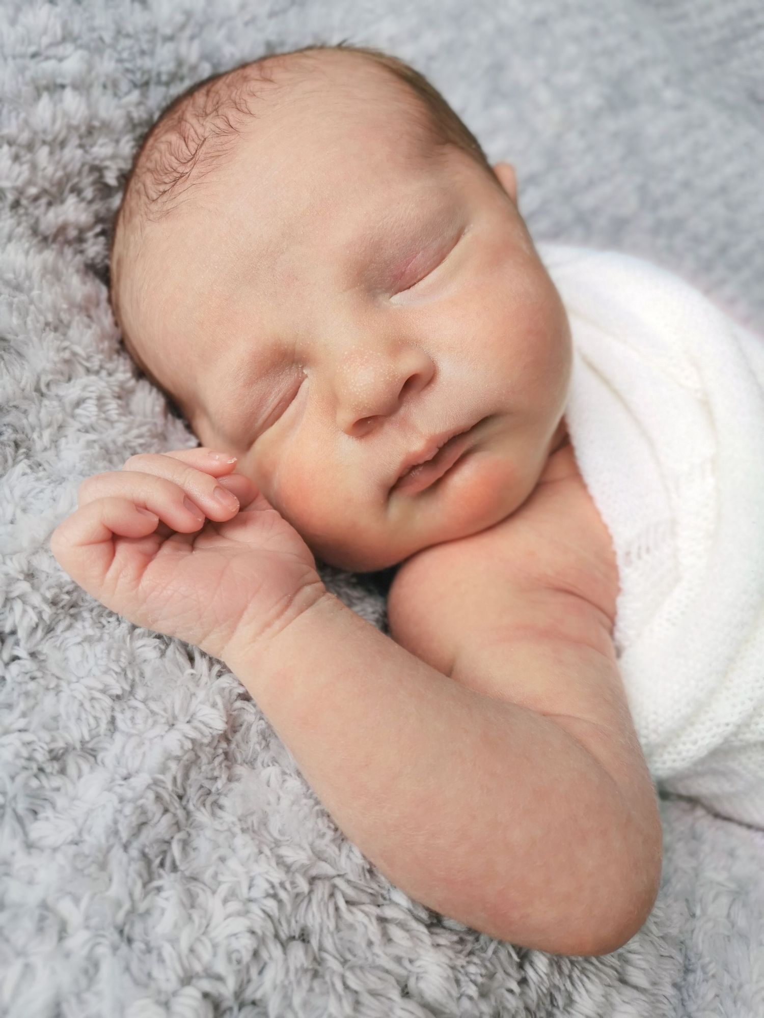 Peaceful newborn baby sleeping on a fluffy gray blanket wrapped in a white swaddle.