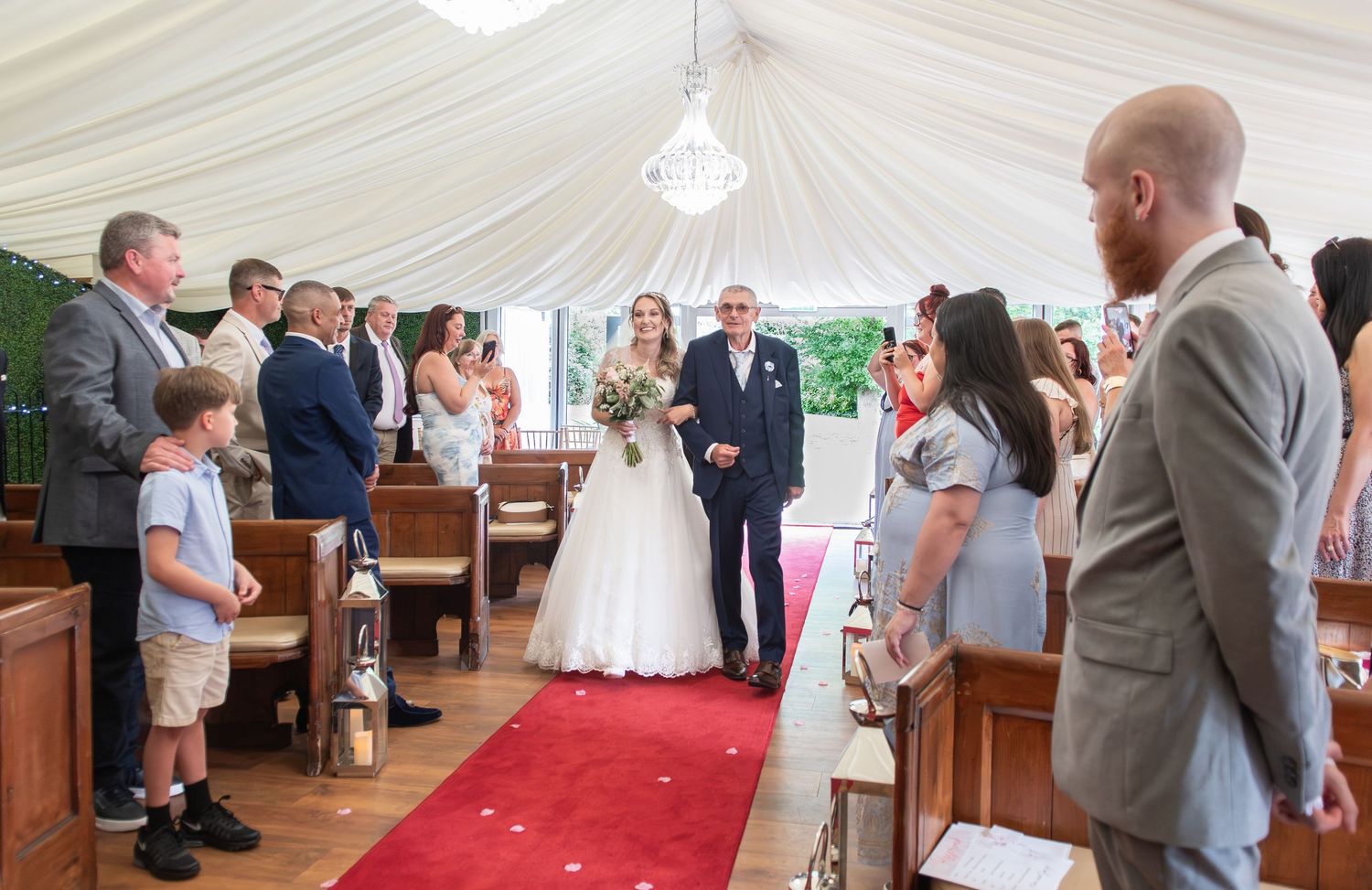 Bride walks down the aisle with her father at a elegant white marquee wedding ceremony with guests standing.