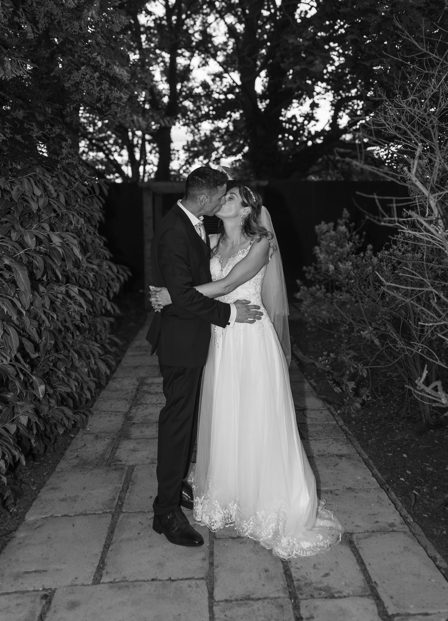 Black and white photo of a bride and groom sharing a romantic kiss on a garden pathway surrounded by lush greenery.