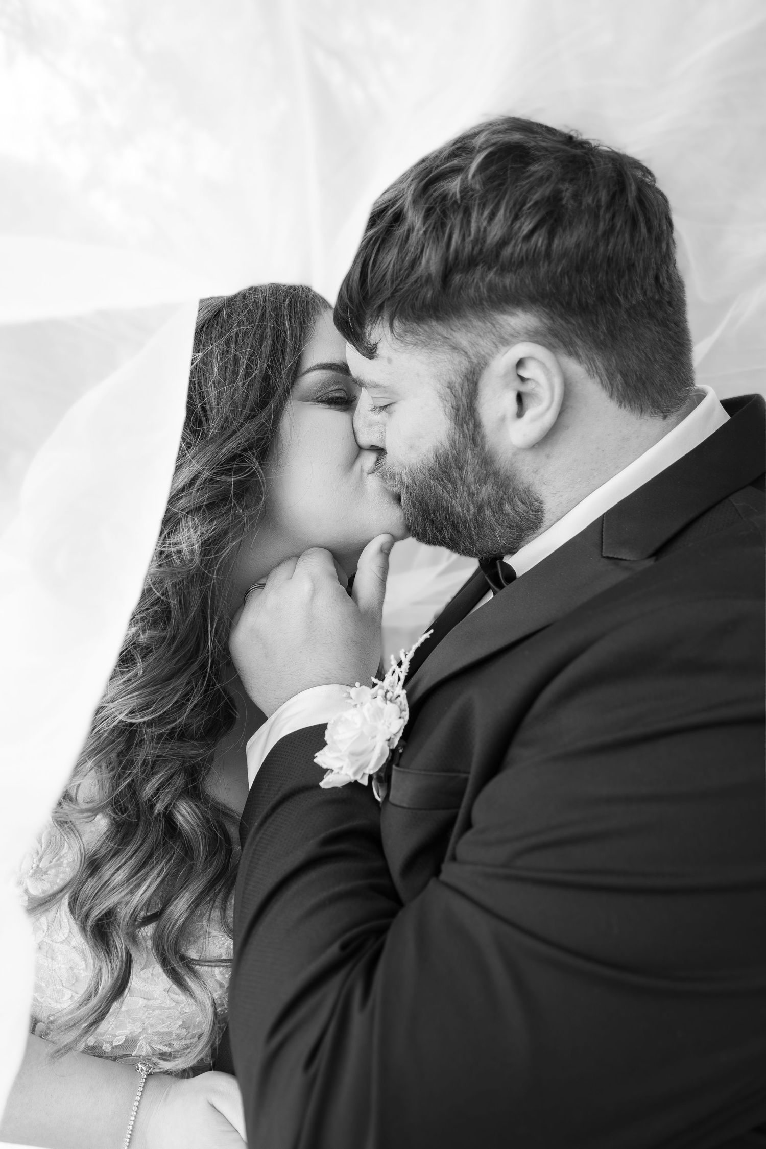 Black and white photo of a bride and groom sharing a romantic kiss under a veil on their wedding day.