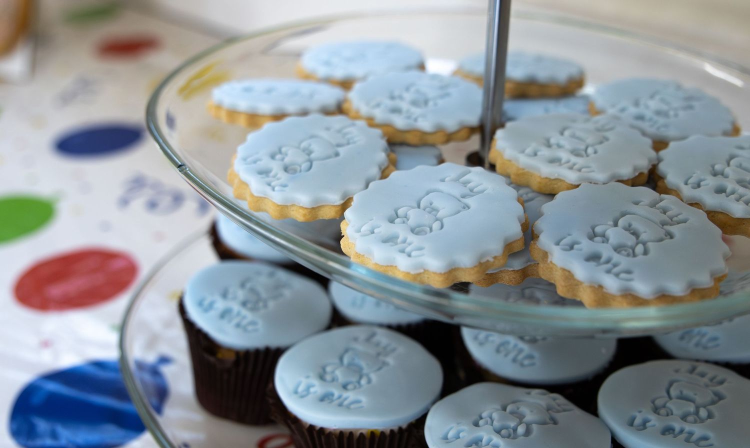 Blue frosted decorated cookies on a two-tier glass cake stand at a baby boy birthday party.