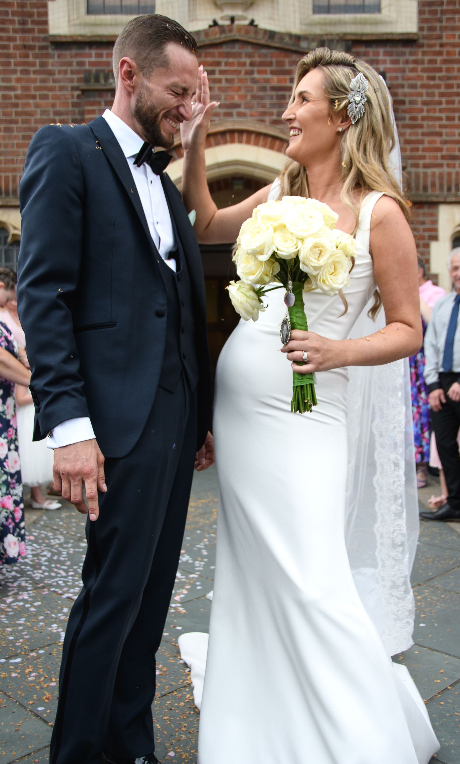 A bride in a fitted white gown holds a yellow bouquet while sharing a joyful moment with her groom outside a brick building.