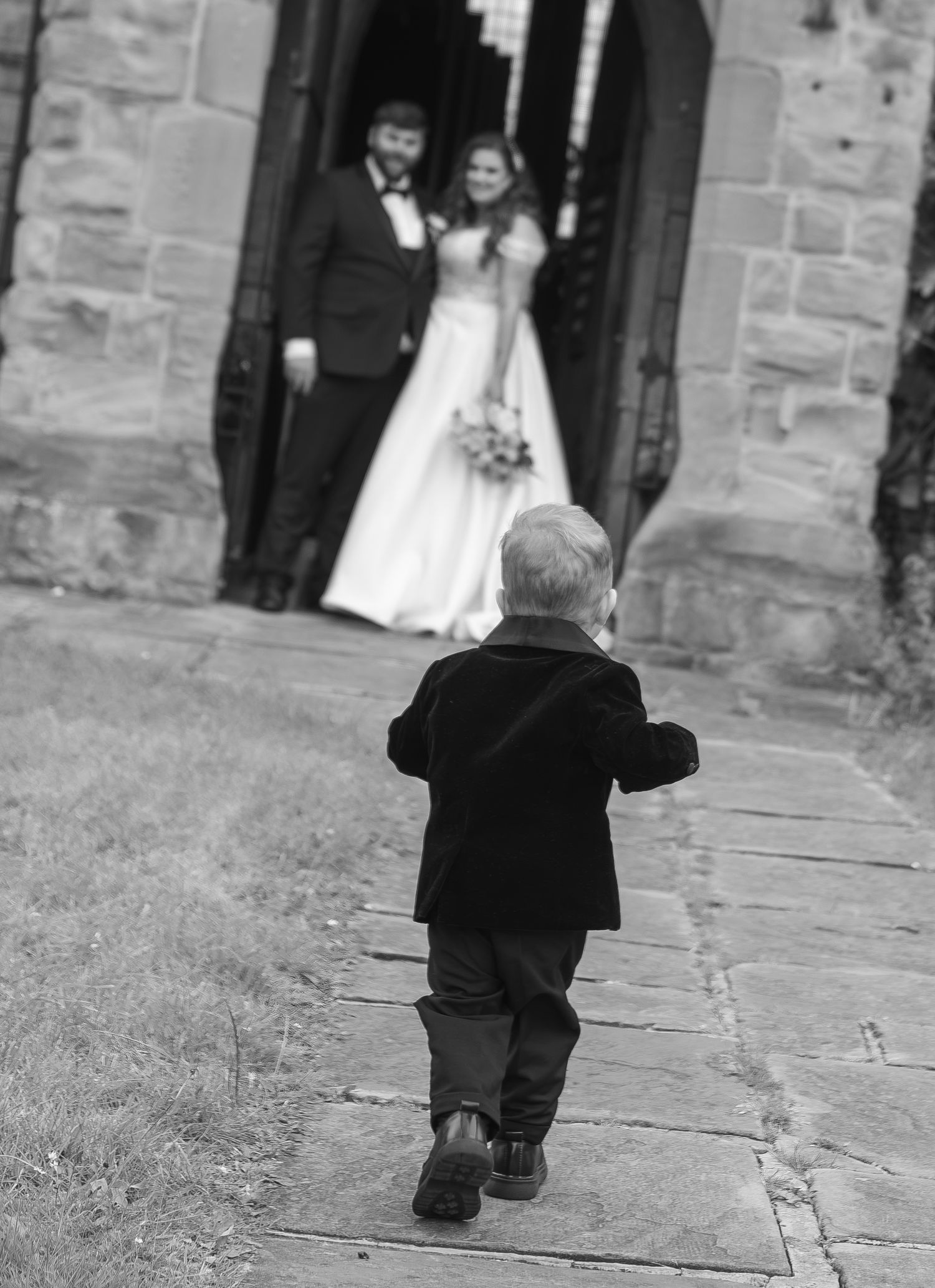 Black and white photo of a toddler in a suit walking toward a bride and groom standing in a church doorway.