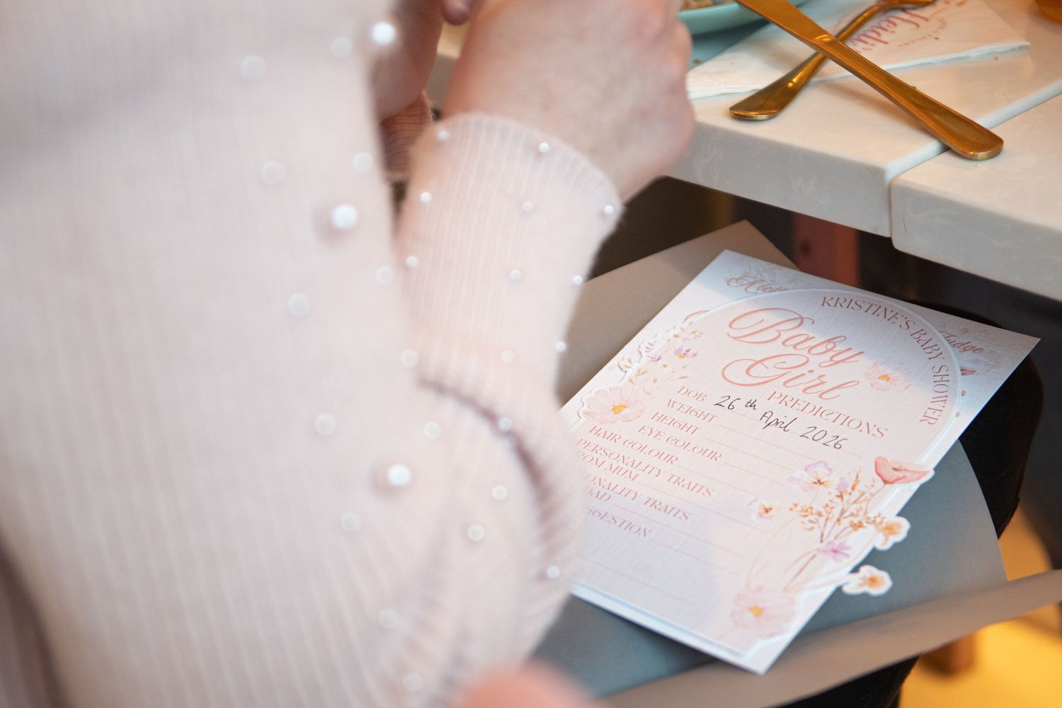 Close-up of a person in a pearl-embellished white sweater holding a floral pink baby shower invitation card.