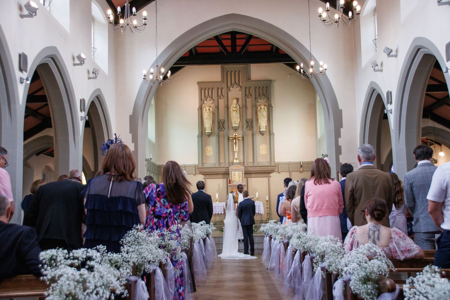Guests stand in a decorated church aisle during a wedding ceremony, with the couple at the altar and floral pew ends.
