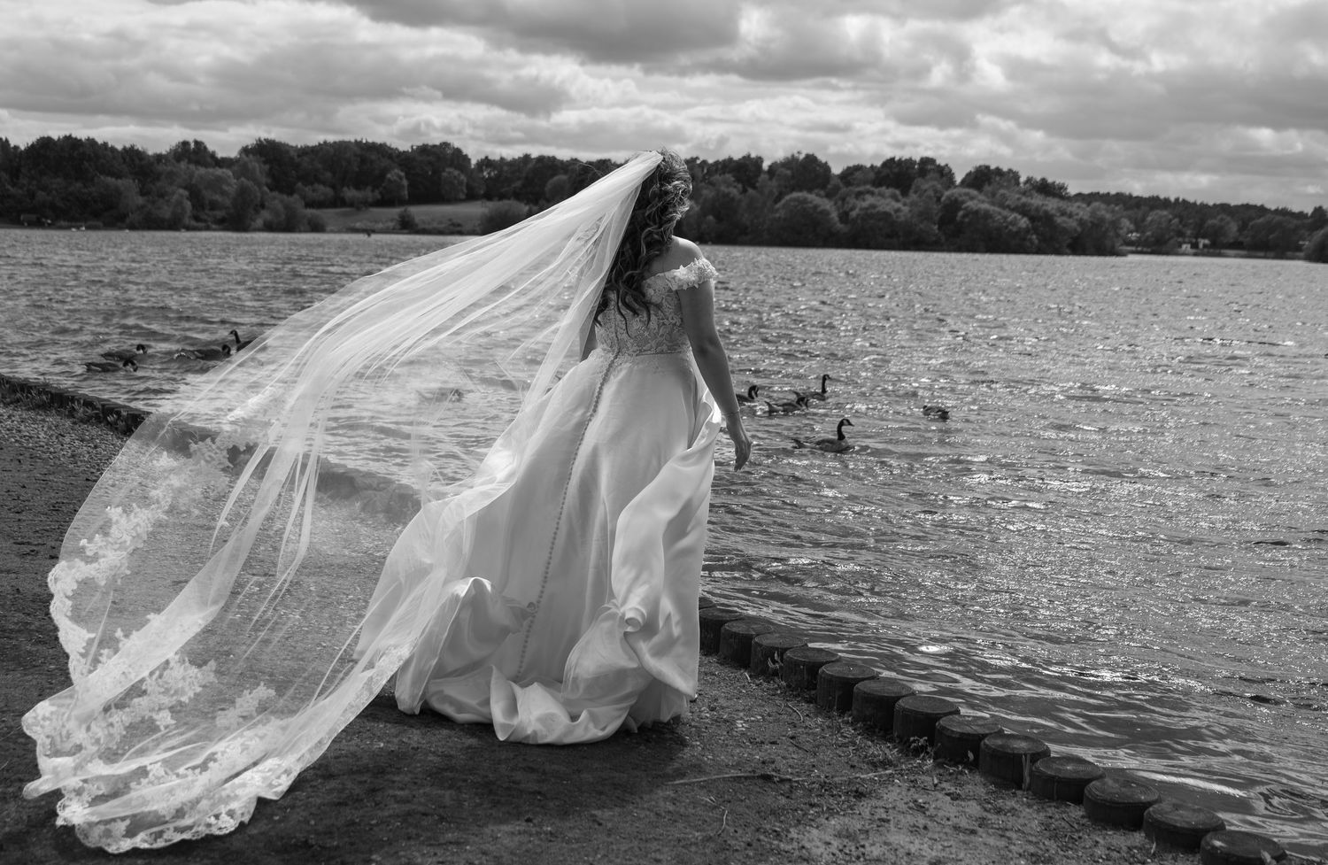 Bride in flowing white gown with veil blowing in wind beside a serene lake in black and white.