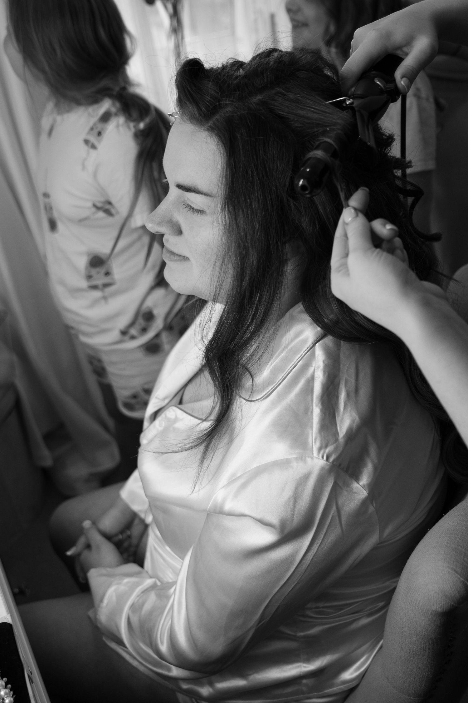 Black and white photo of a woman in a satin robe getting her hair curled by a stylist before a wedding.