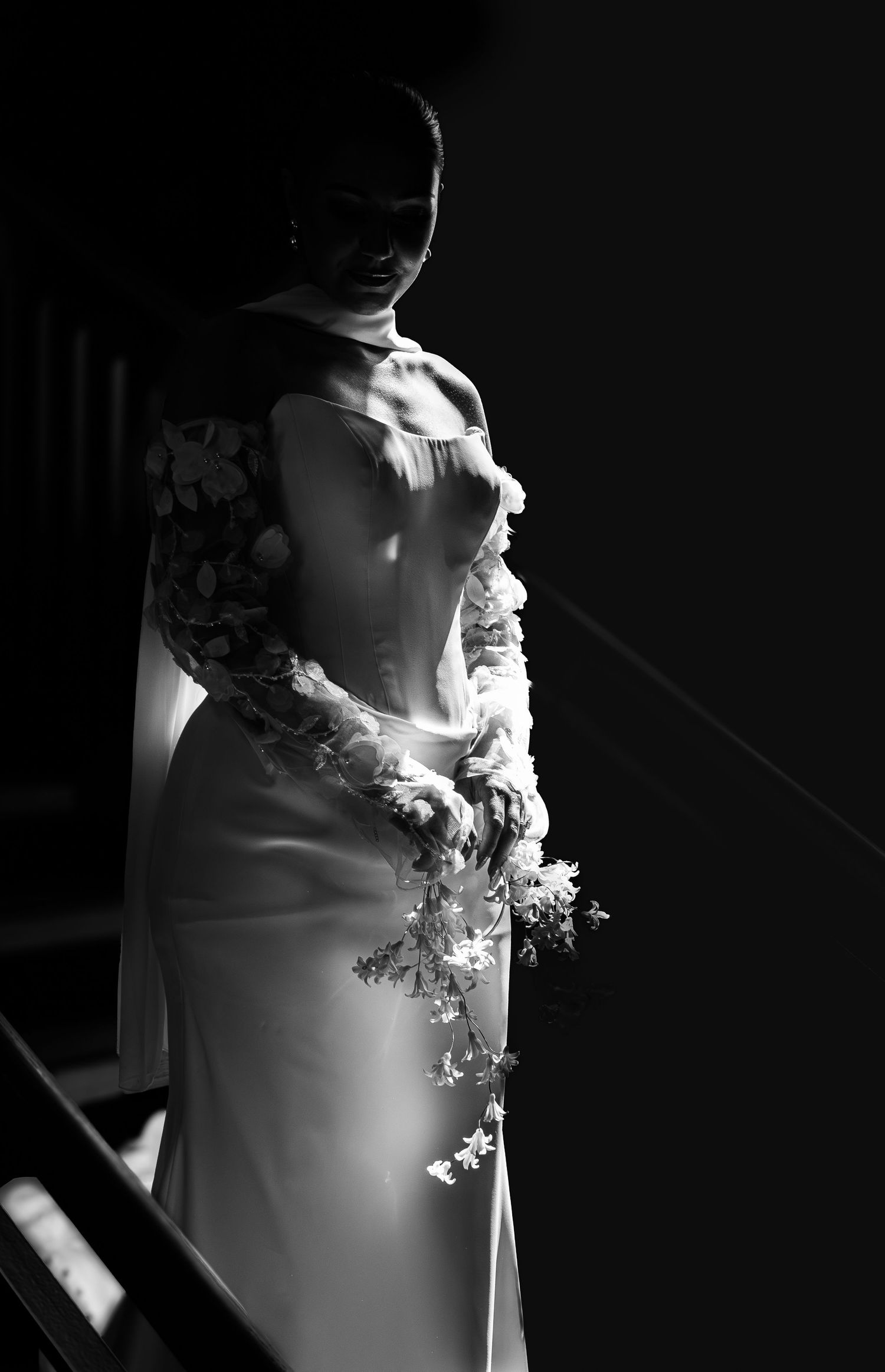 Bride in elegant white gown holding floral bouquet, dramatic black and white wedding portrait.