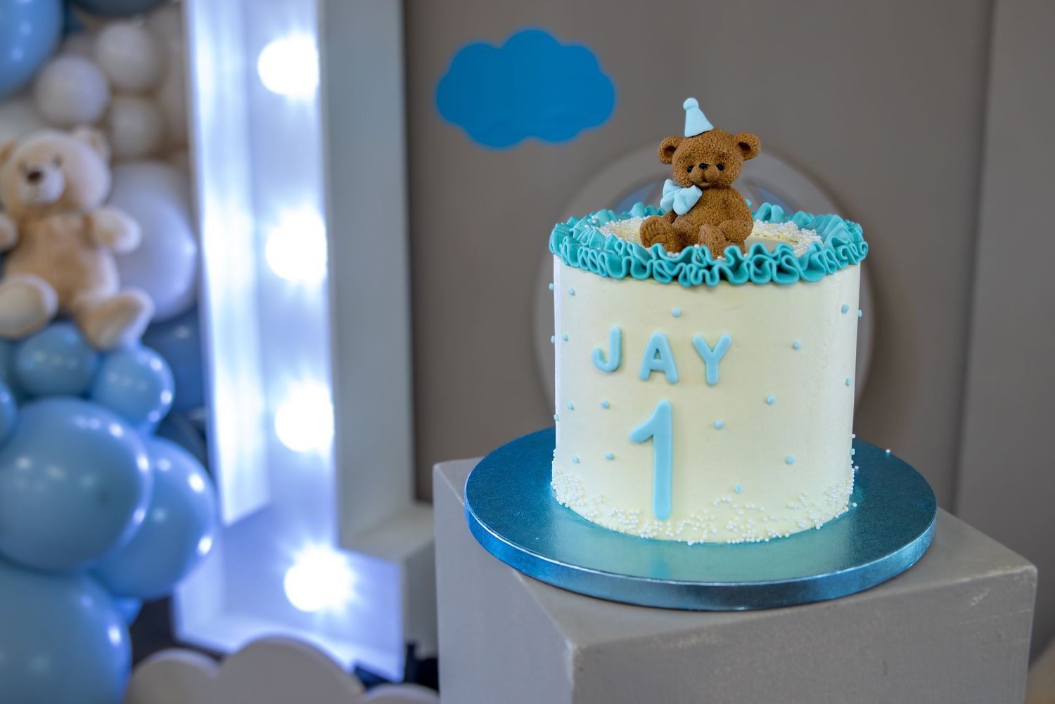 White first birthday cake for Jay decorated with blue frosting, a teddy bear topper, and the number 1, surrounded by blue balloons.
