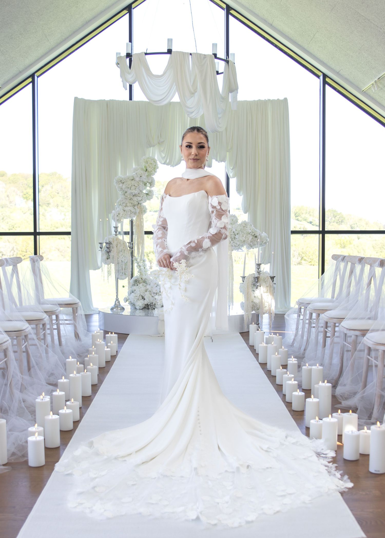 Bride in stunning white off-shoulder gown with cape walks down candlelit aisle in elegant chapel venue.