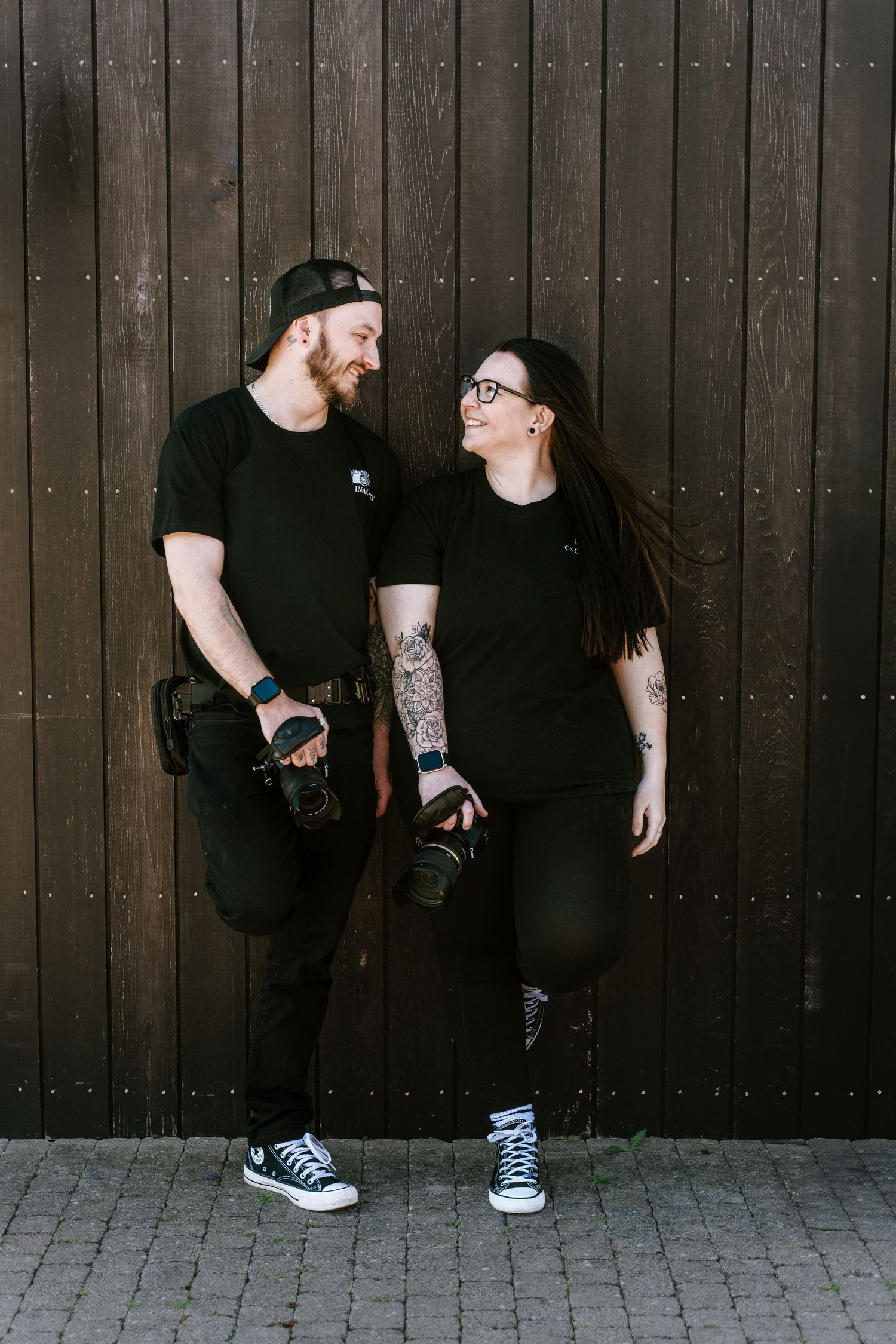 A tattooed couple in matching black outfits holding cameras, smiling at each other against a dark wooden wall.