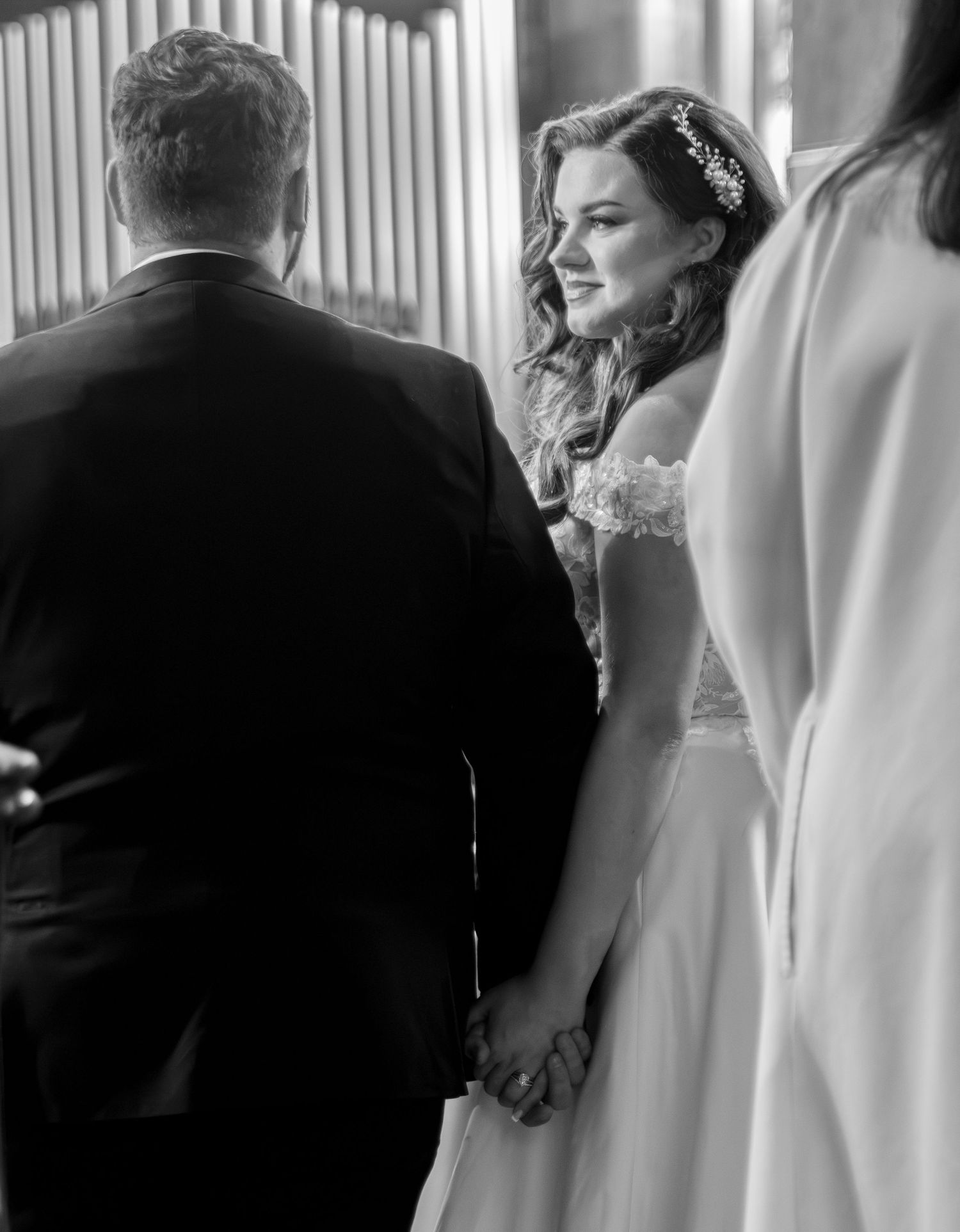 Black and white wedding photo of bride holding groom's hand at altar, wearing off-shoulder gown with hair accessory.