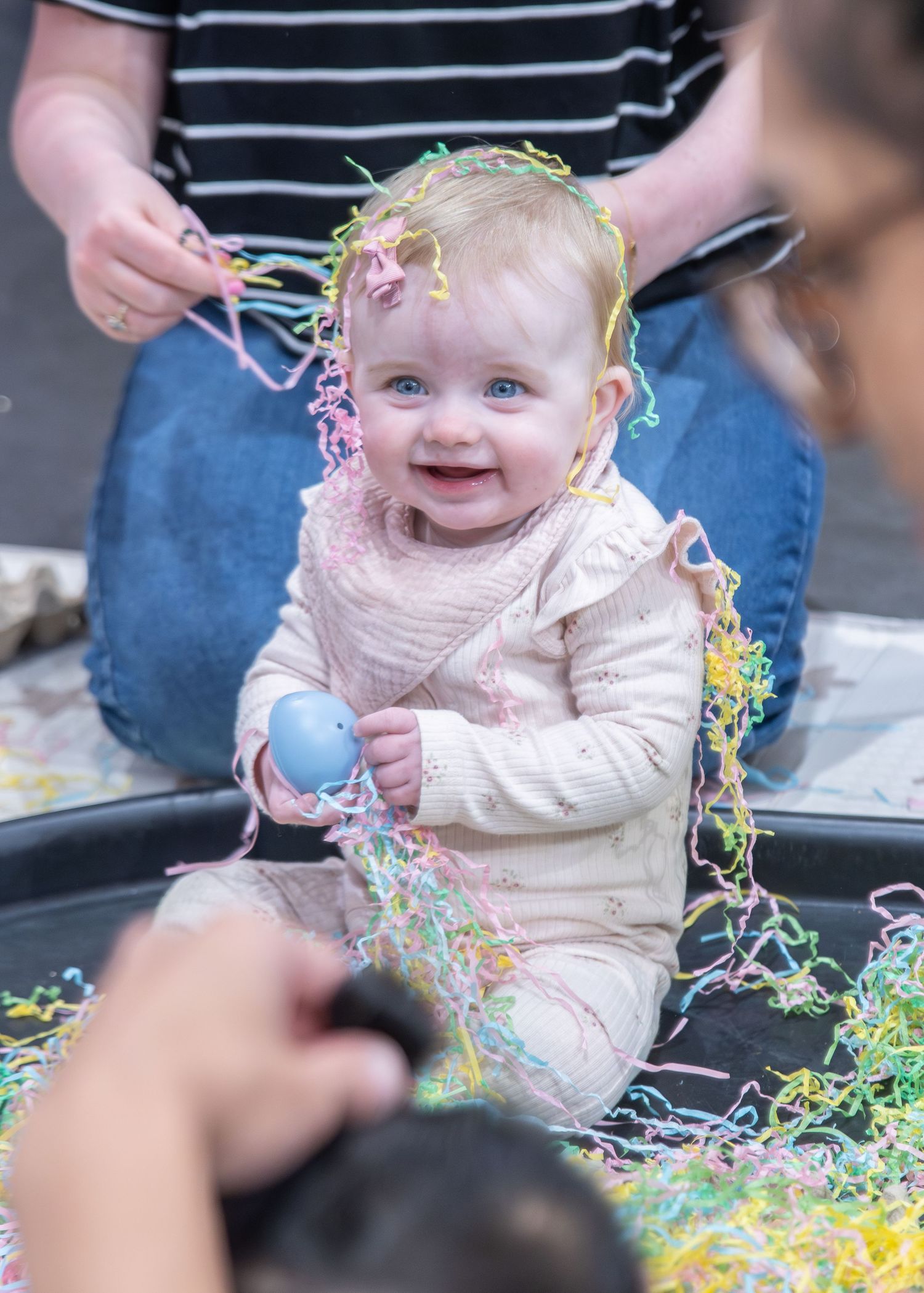 Smiling baby with blue eyes holds Easter egg surrounded by colorful paper shreds during festive Easter celebration.