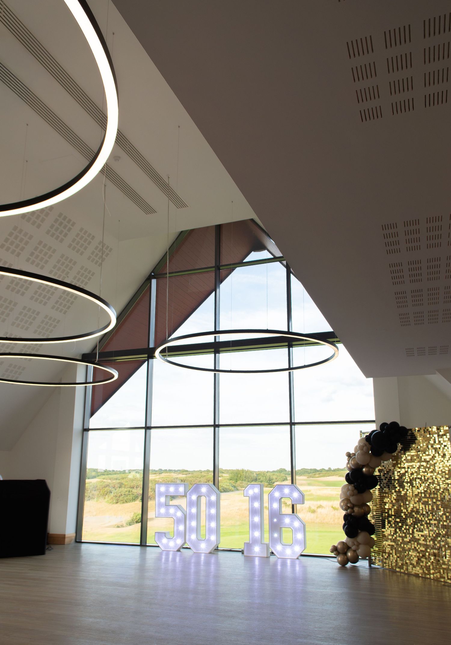 Modern event venue with illuminated 50 & 16 marquee numbers, balloon decorations, circular pendant lights, and floor-to-ceiling windows.