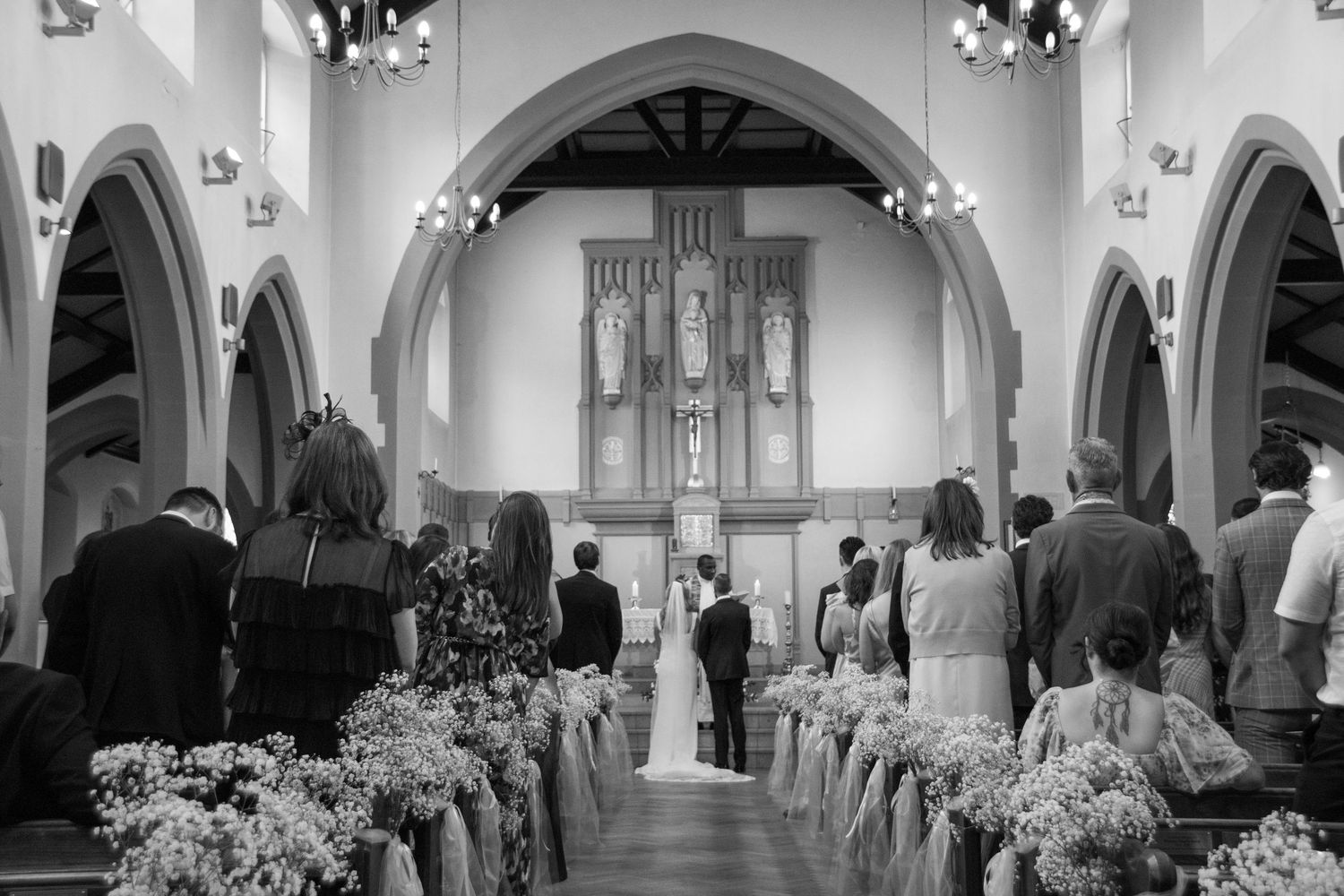 Black and white photograph of a wedding ceremony inside a church with arched architecture and floral decorations along the aisle.