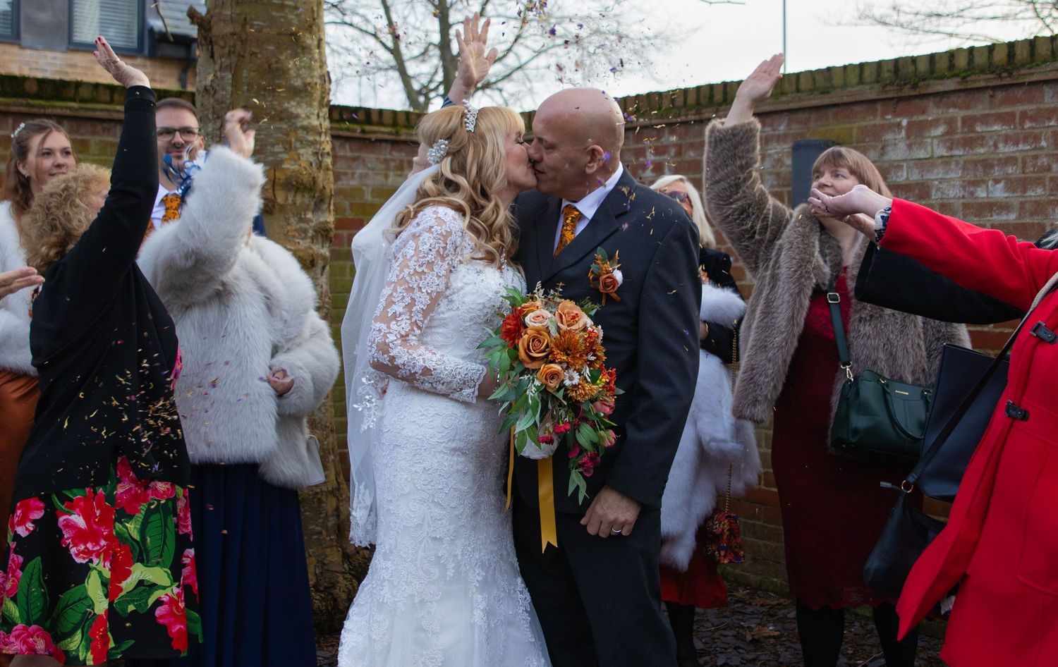 Wedding guests shower newlyweds with confetti during outdoor celebration by brick wall.