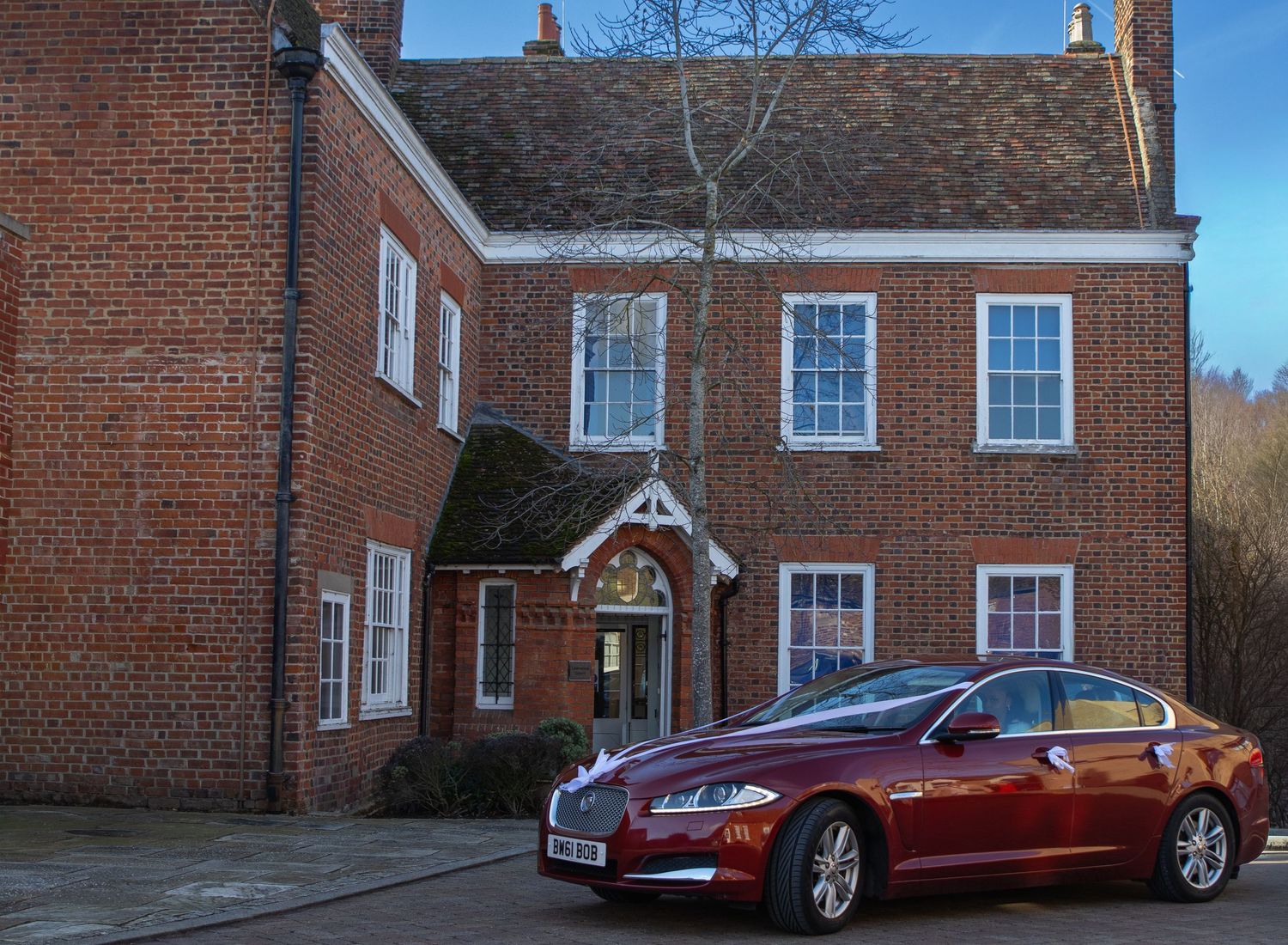 Red luxury car parked in front of traditional brick Georgian-style house on sunny day.