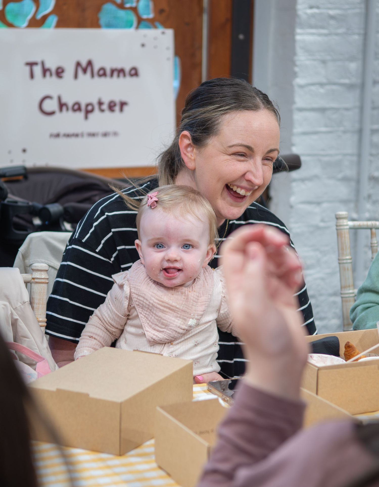 A laughing mother holds her smiling baby girl at The Mama Chapter event, surrounded by boxes on a table.
