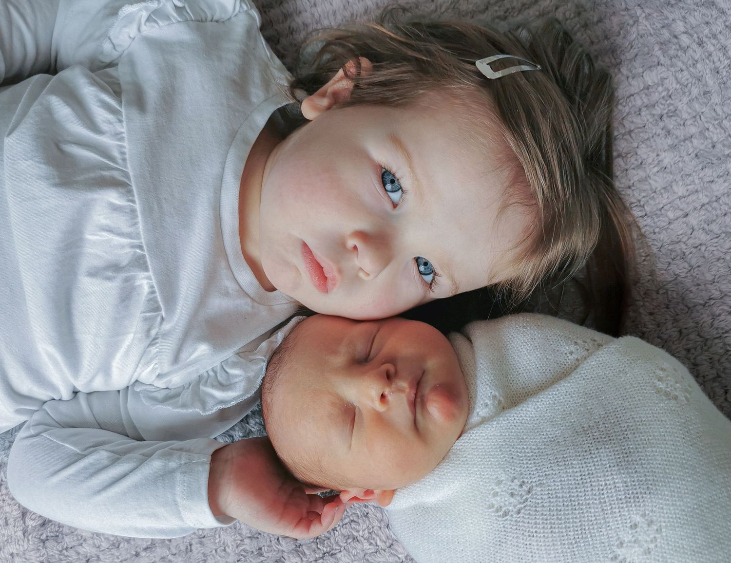 Toddler girl with blue eyes lying beside a sleeping newborn baby wrapped in a white swaddle.