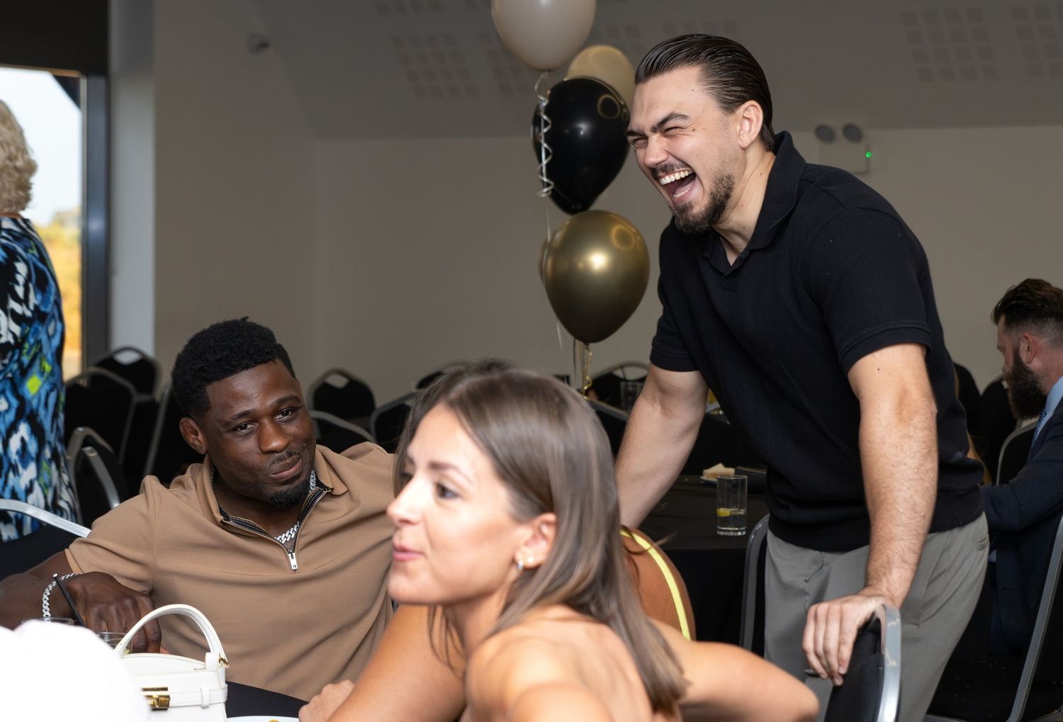 Adults laughing and socializing at a party with black and gold balloon decorations.