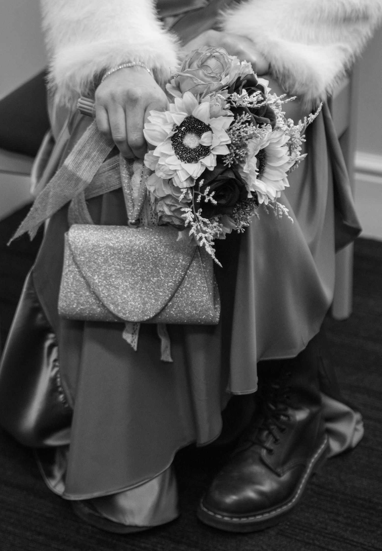 Black and white photo of bride holding sunflower bouquet and glitter clutch, wearing satin skirt and lace-up boots.