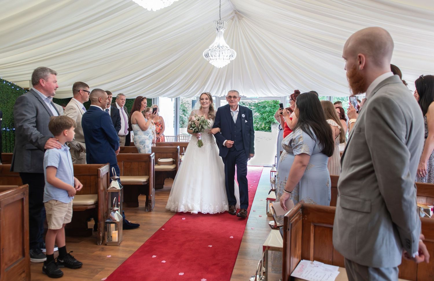 Bride walks down the aisle with her father at an elegant marquee wedding ceremony with guests standing.