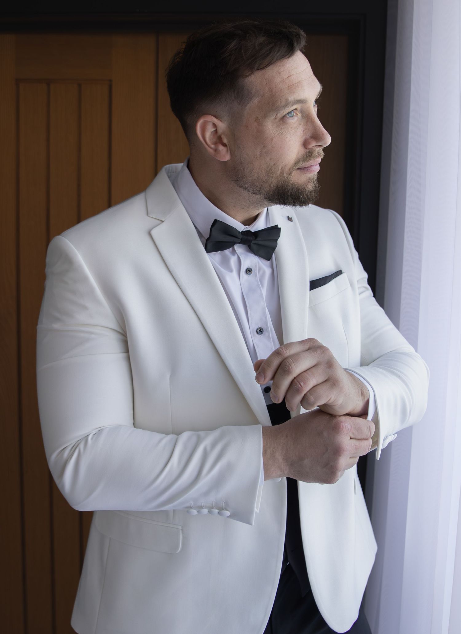 Handsome groom in white tuxedo jacket and black bow tie adjusting cufflinks by window on wedding day.