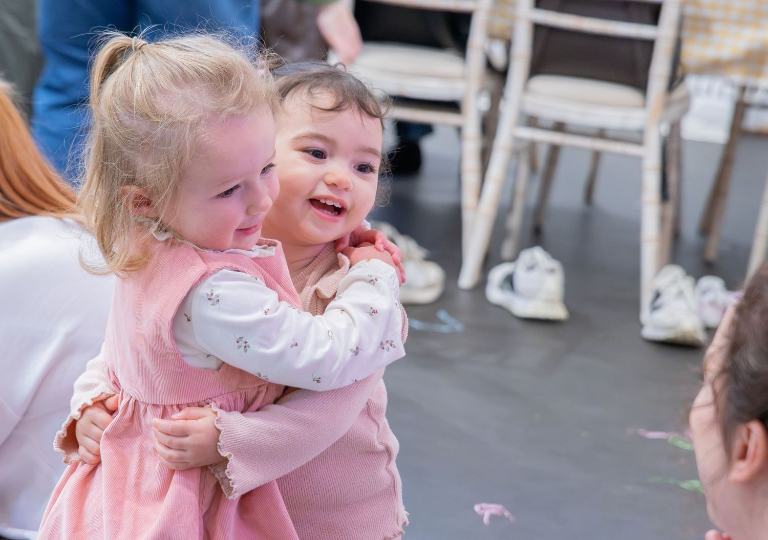 Two toddlers sharing a joyful hug at an indoor event, both smiling brightly in adorable outfits.