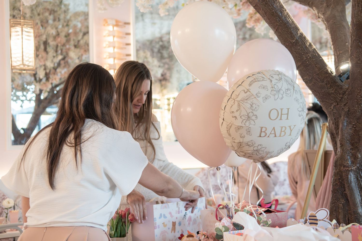 Two women arranging decorations at a pink baby shower with OH BABY balloons and floral table settings.