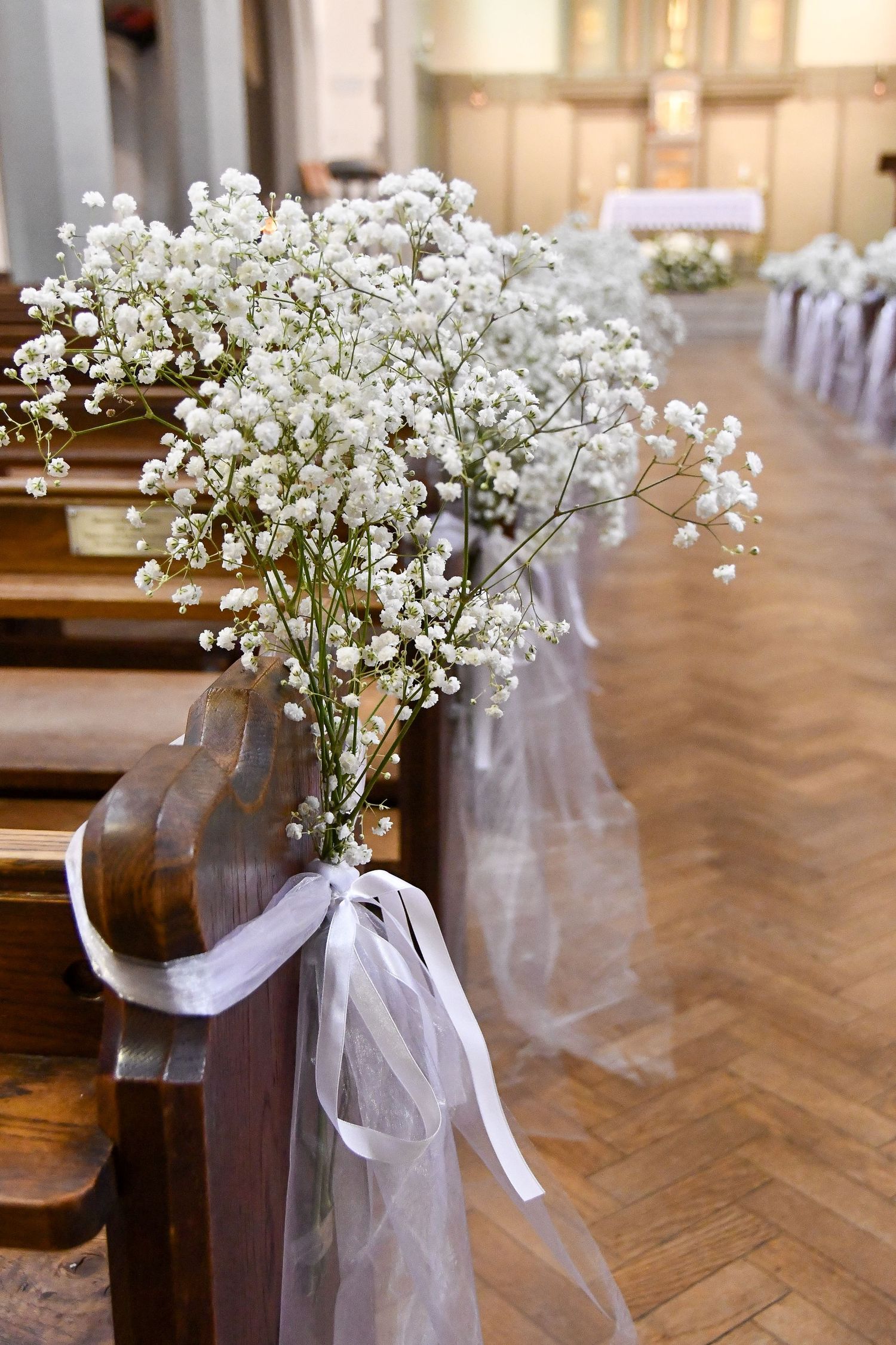 Church pews decorated with baby's breath flowers and white tulle bows along a herringbone wood floor.