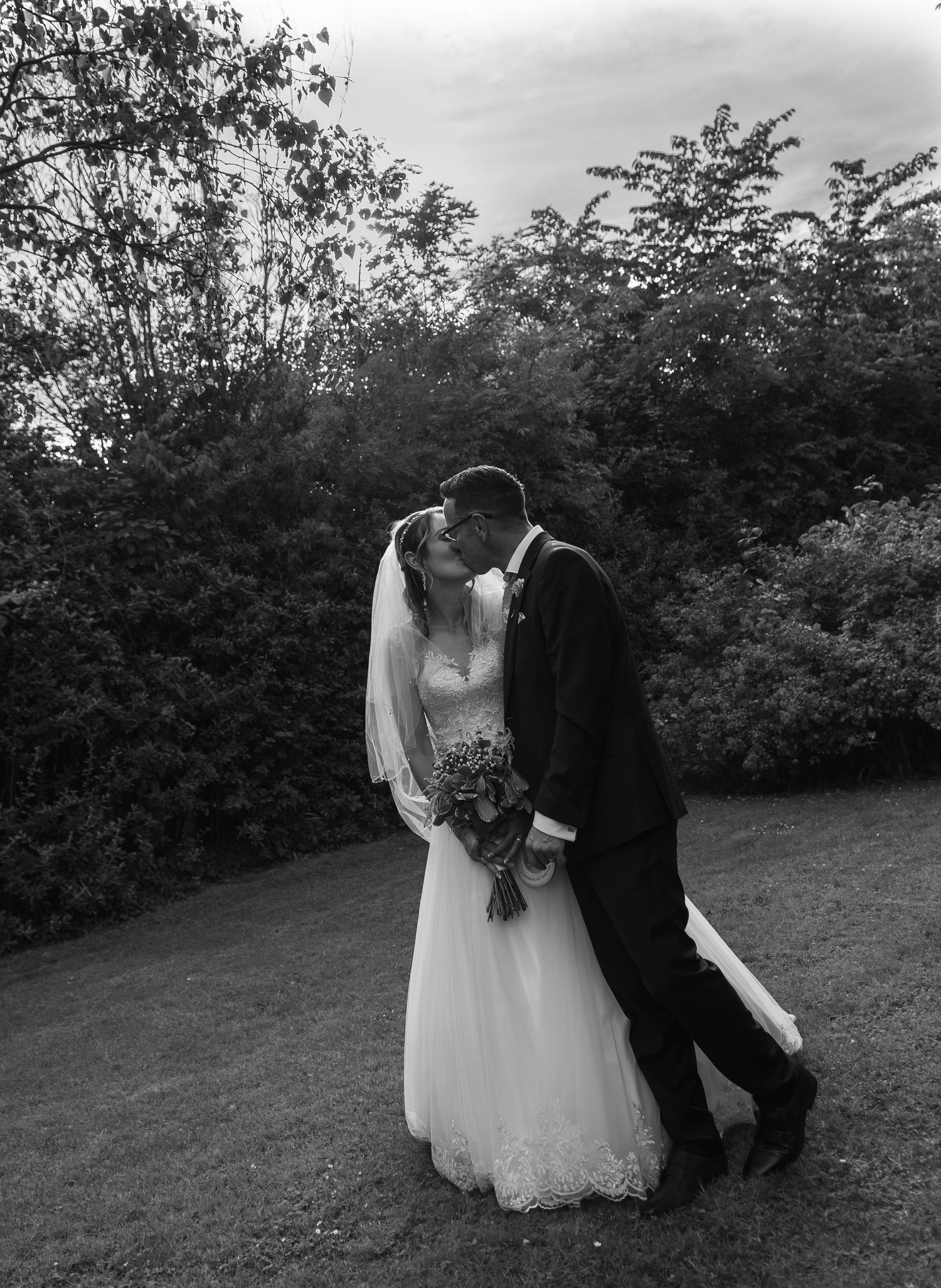 Black and white photo of a bride and groom sharing a kiss outdoors, surrounded by lush green trees on their wedding day.
