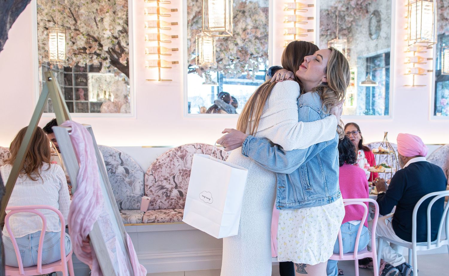 Two women hugging warmly inside a chic pink cafe, one holding a white shopping bag.