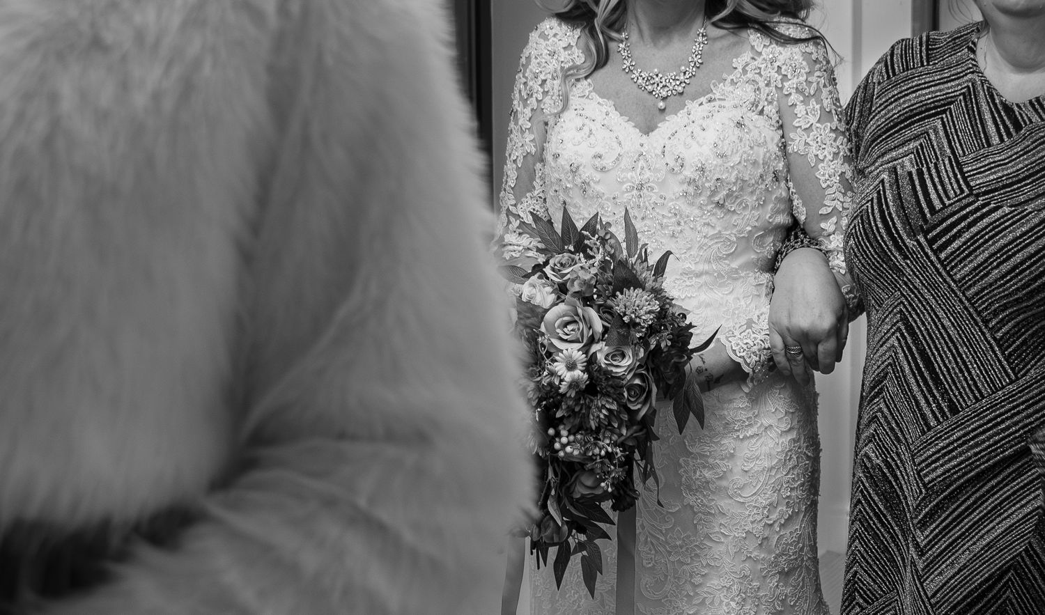 Black and white photo of a bride in lace gown holding a floral bouquet, standing beside a woman in a striped dress.