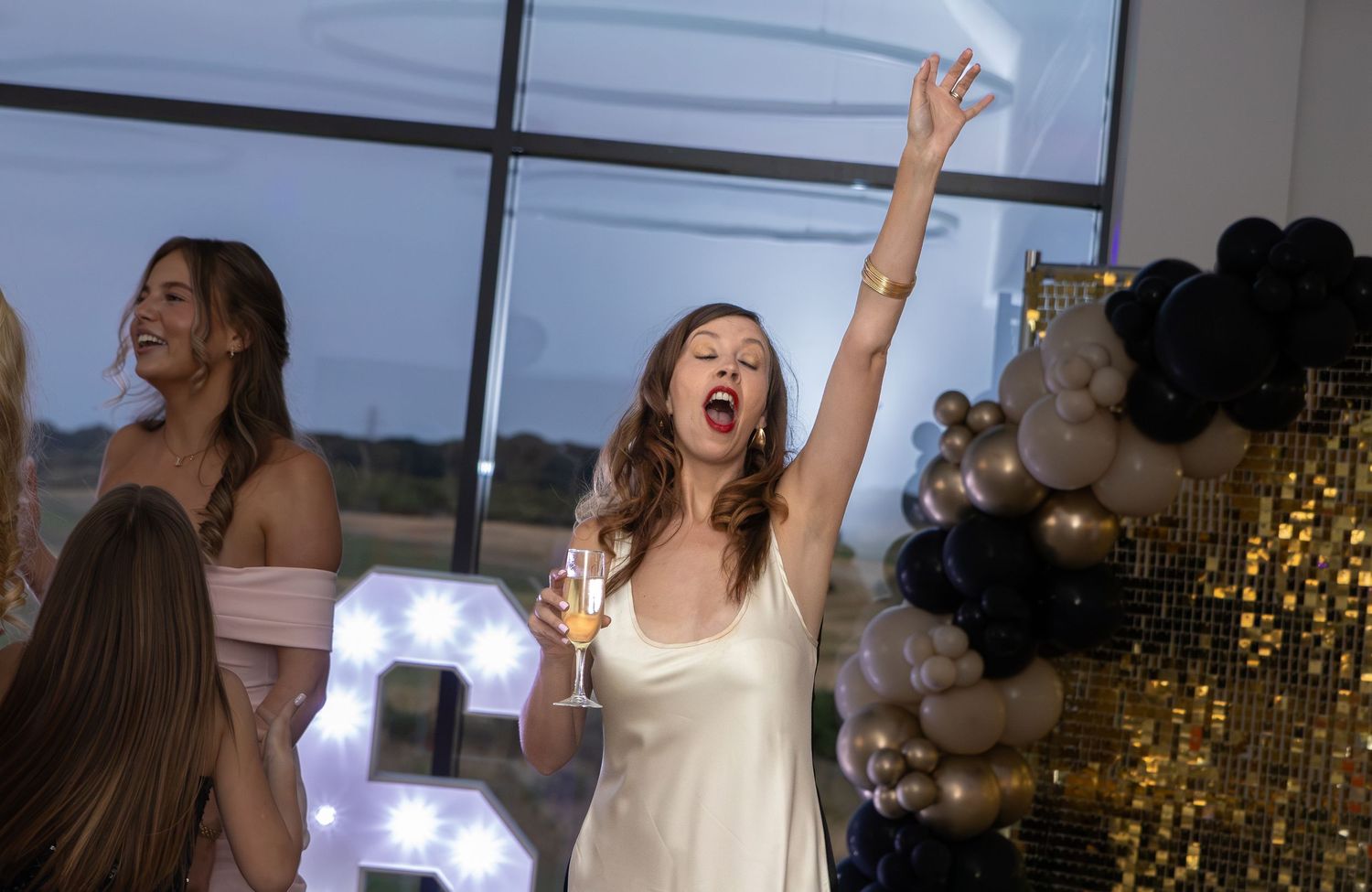 Woman in white dress celebrating with champagne at a party with balloons and decorative letter backdrop.