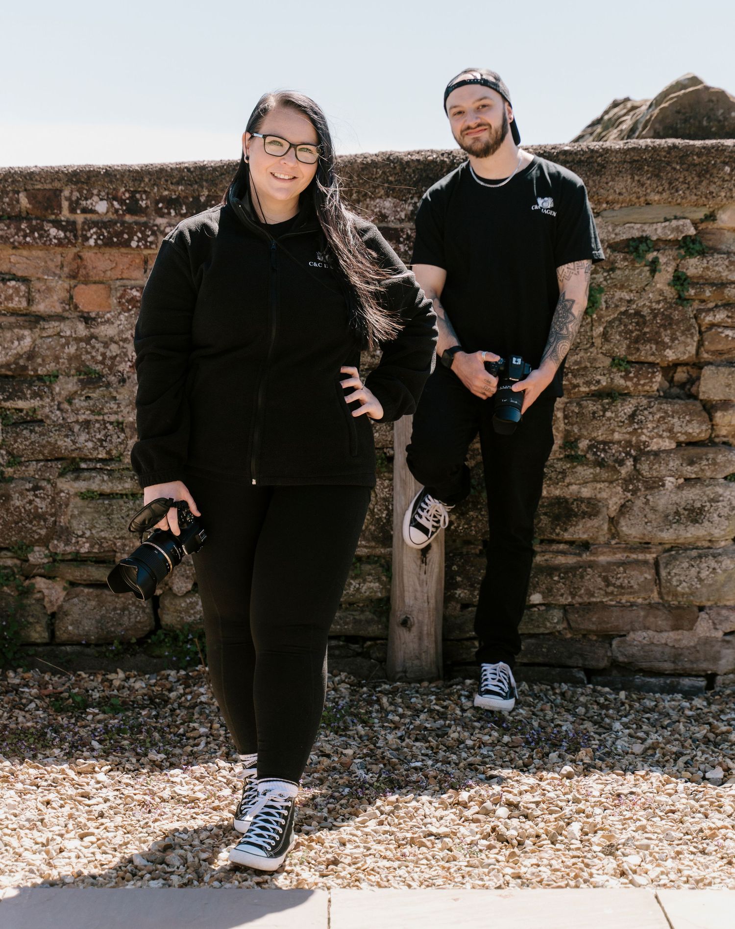 Two photographers in black outfits holding cameras pose against a rustic stone wall outdoors on a sunny day.