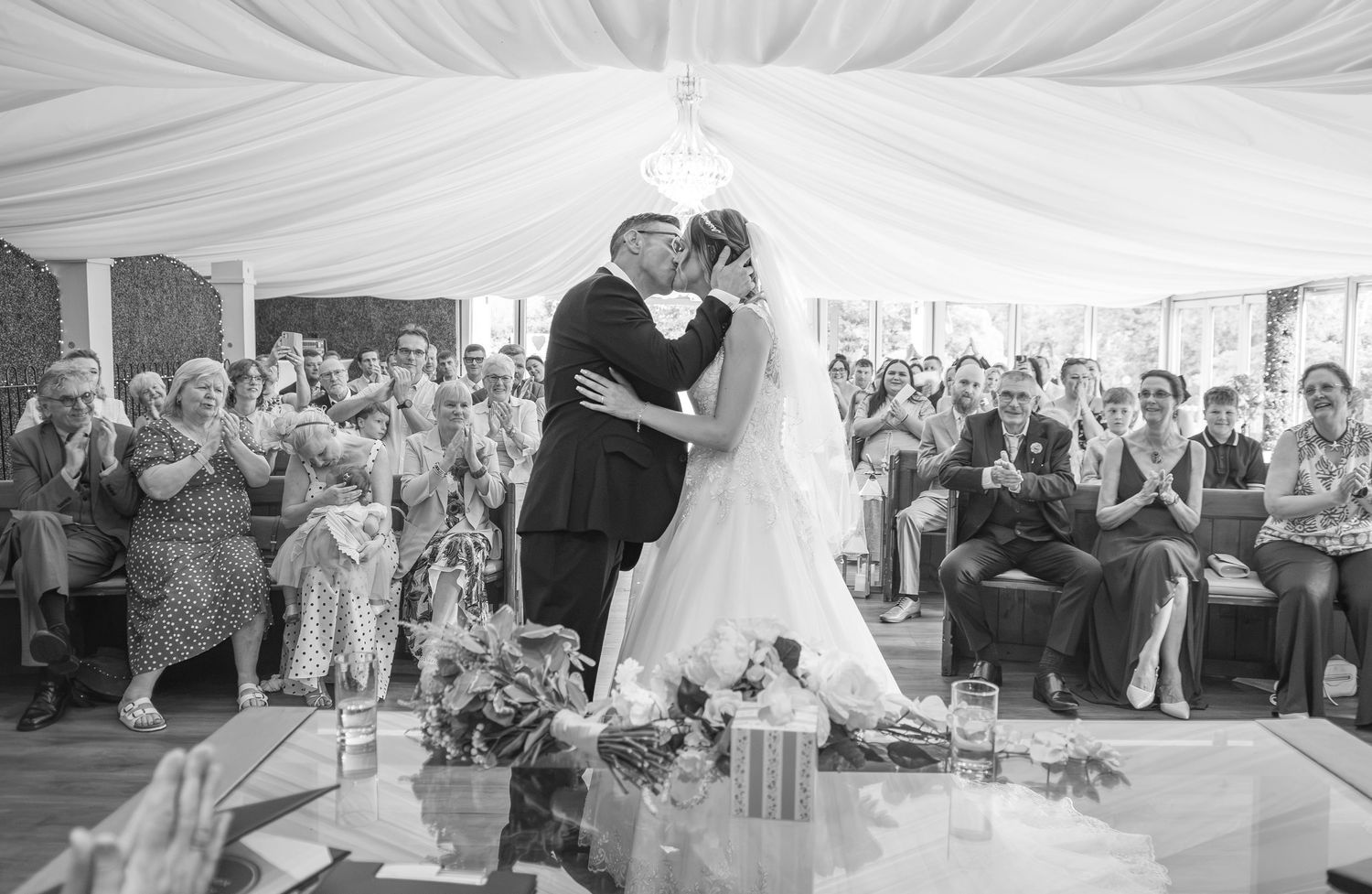 Black and white photo of a bride and groom sharing their first kiss at the altar, surrounded by applauding wedding guests.