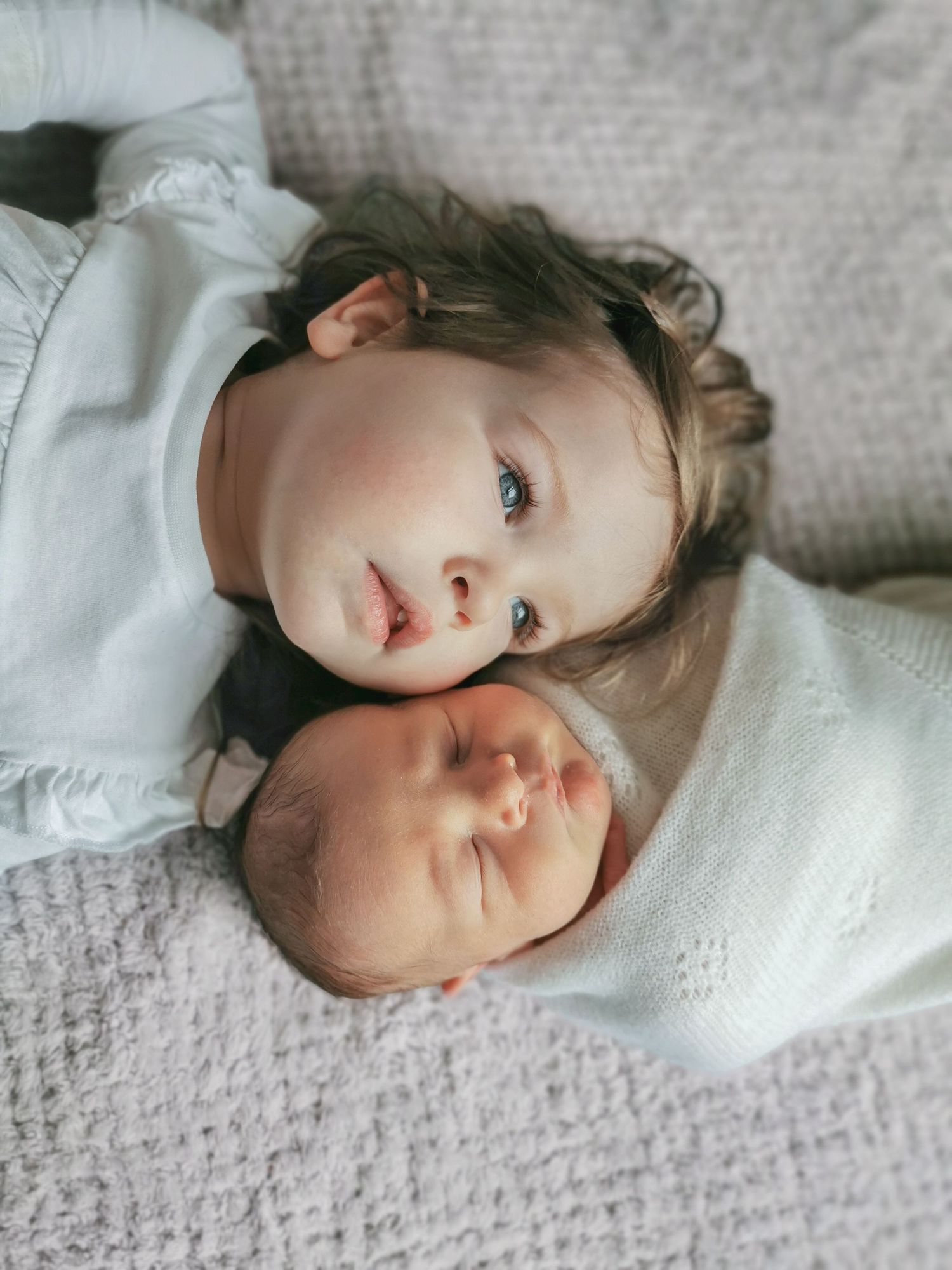 Toddler girl with blue eyes lying beside sleeping newborn baby sibling on white blanket.