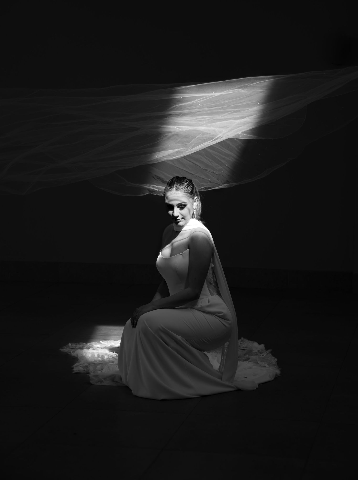 Elegant woman in gray gown kneeling under flowing veil in dramatic black and white portrait photography.