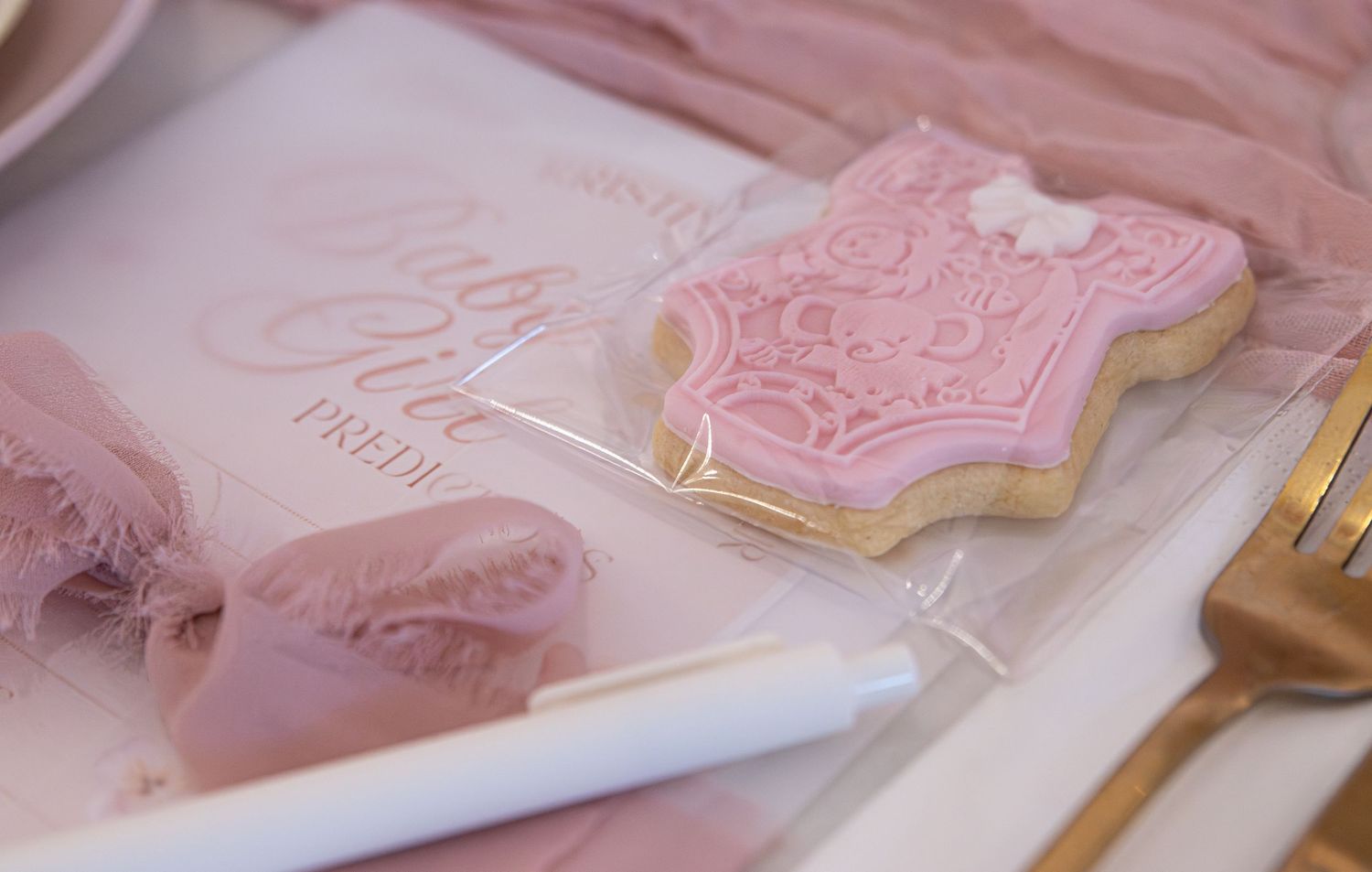 Pink decorated sugar cookie in clear bag beside a gold fork and pink paper at an elegant baby shower table setting.
