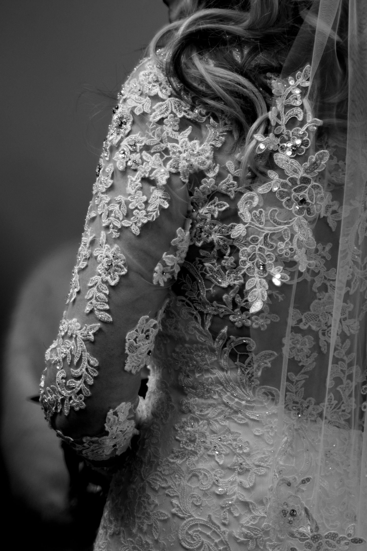 Black and white close-up of a bride in an intricate lace wedding gown with long sleeves and delicate floral embroidery.