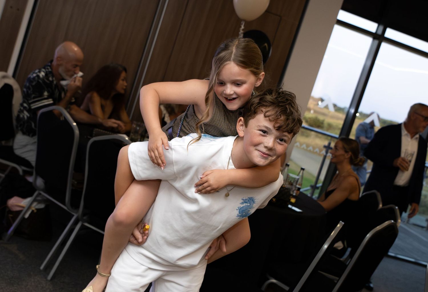 Young boy giving a smiling girl a piggyback ride at an indoor celebration event.