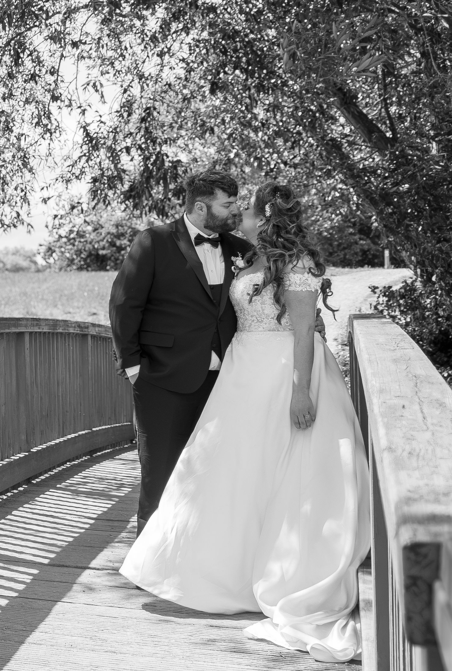 Black and white photo of a bride and groom sharing a kiss on a wooden bridge surrounded by lush trees.