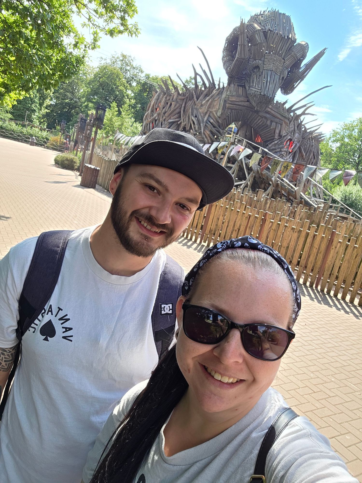 A couple takes selfies at an amusement park with wooden structures in the background.