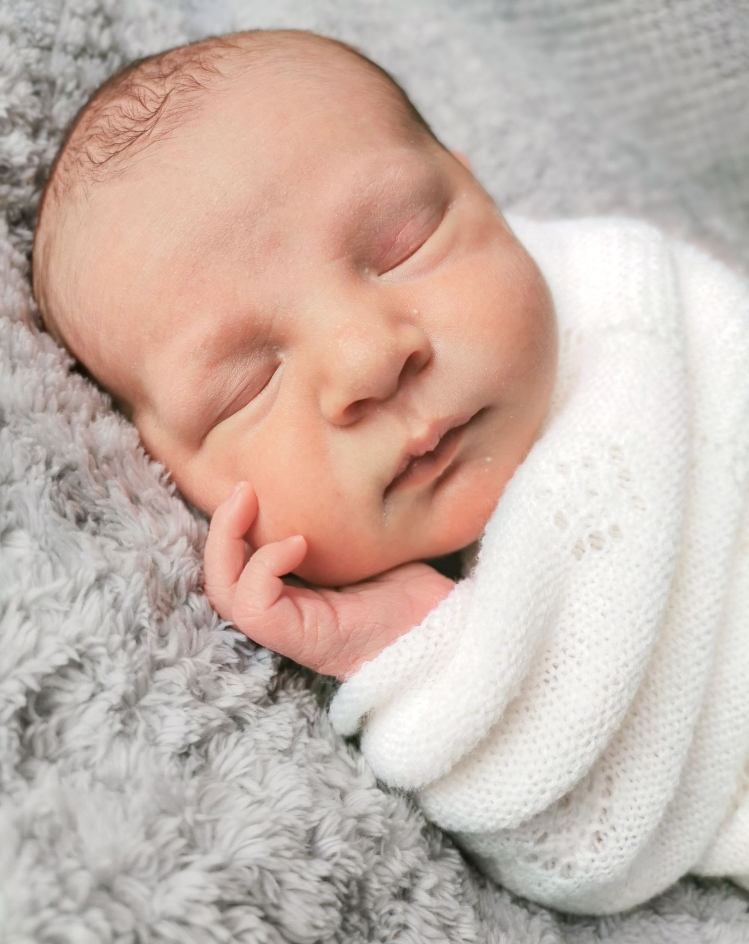 Peaceful newborn baby sleeping swaddled in white knit wrap on soft gray fluffy blanket.