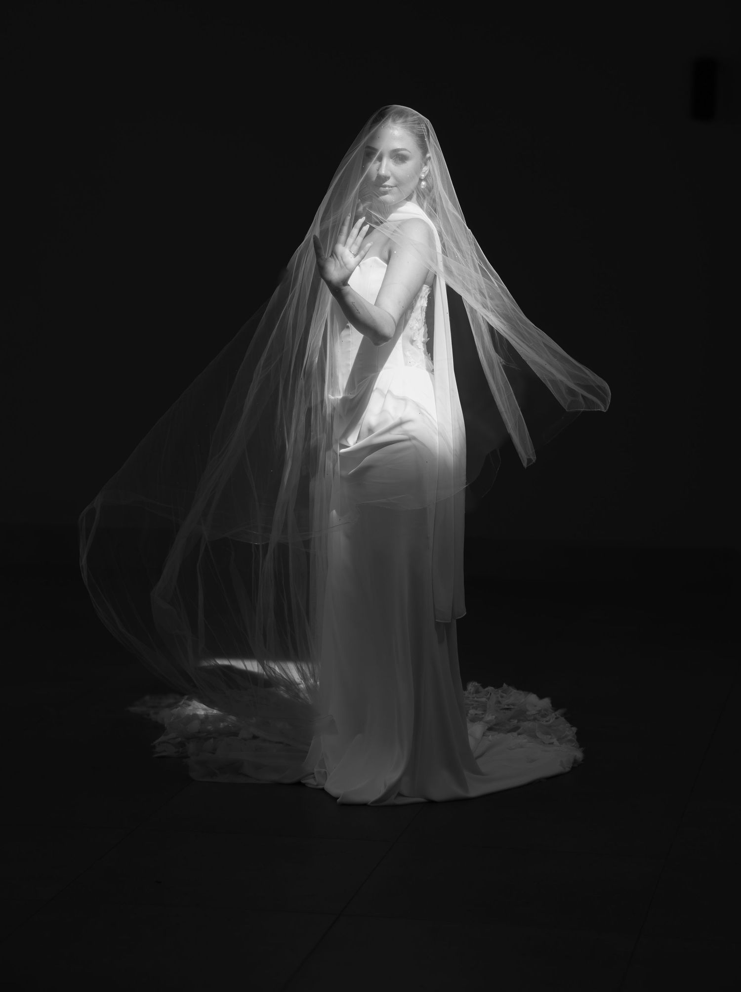 Bride in elegant white wedding gown and long flowing veil poses gracefully against dark background in black and white photo.