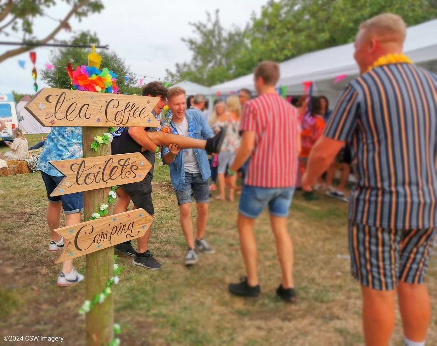 Wooden directional signs at an outdoor event with people mingling in the background.