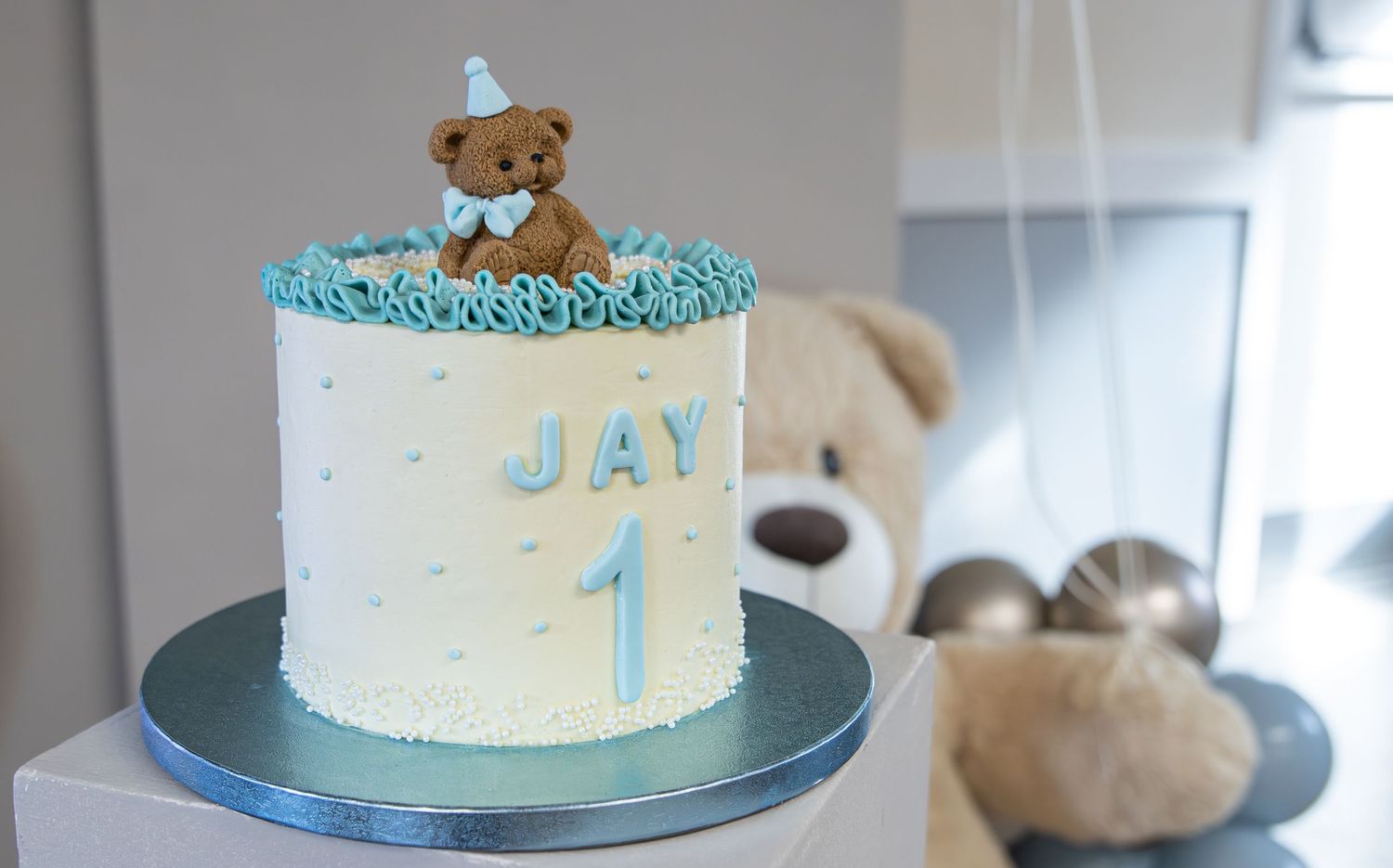 Blue and white first birthday cake with teddy bear topper and name JAY, decorated with rosettes and number 1.