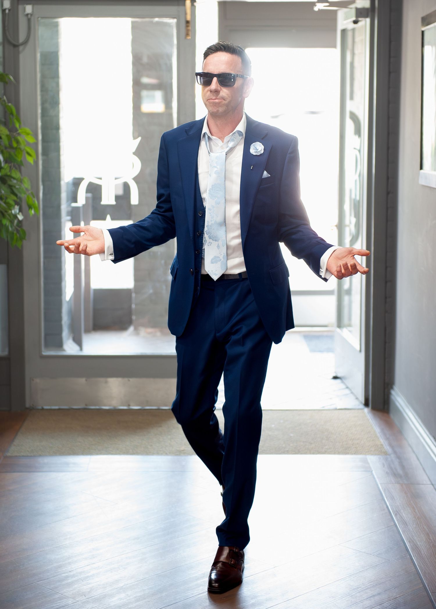 Man in navy blue suit and sunglasses striking a confident pose in a modern hallway.